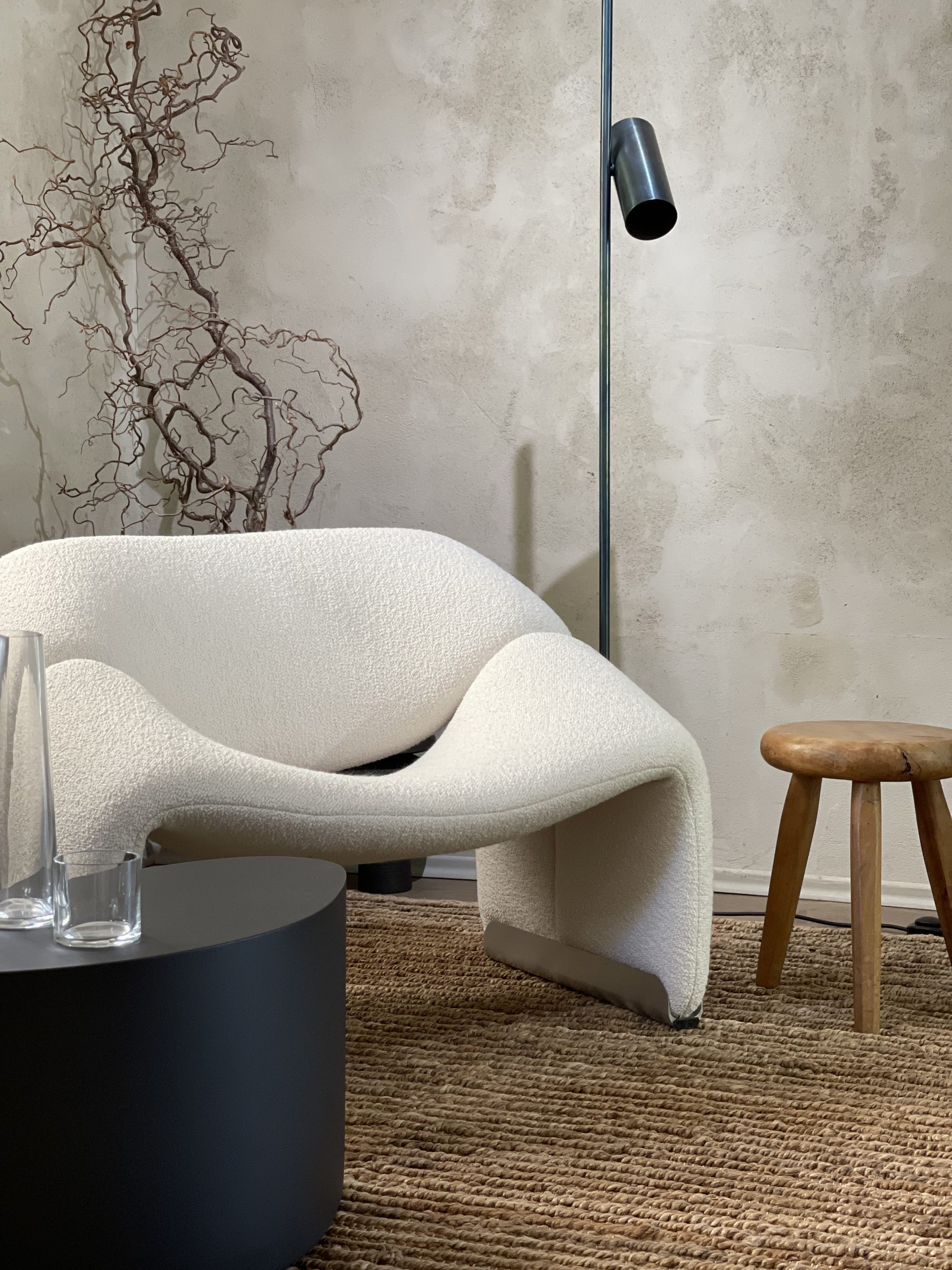 Design-Loungechair Groovy Chair von Pierre Paulin von Artifort mit weißem Bouclé-Bezug vor gespachtelter Kalkputzwand, kombiniert mit Naturteppich, Holzhocker, schwarzem Couchtisch und minimalistischer Stehleuchte – modernes Interior mit organischer Formsprache