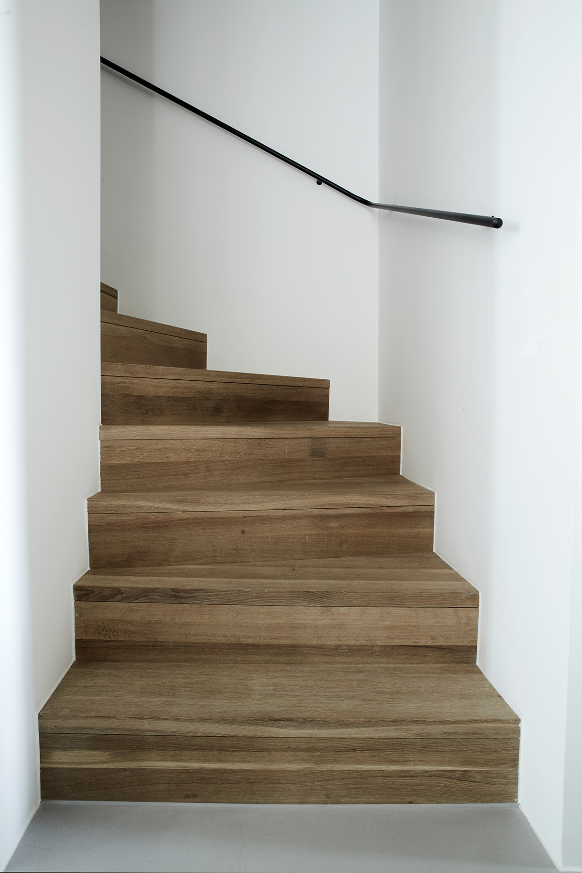 Minimalistische Holztreppe mit hell geöltem Eichenbelag, weiß verputzten Wänden und schlichtem schwarzem Handlauf – moderne Innenarchitektur in klarer Formensprache.