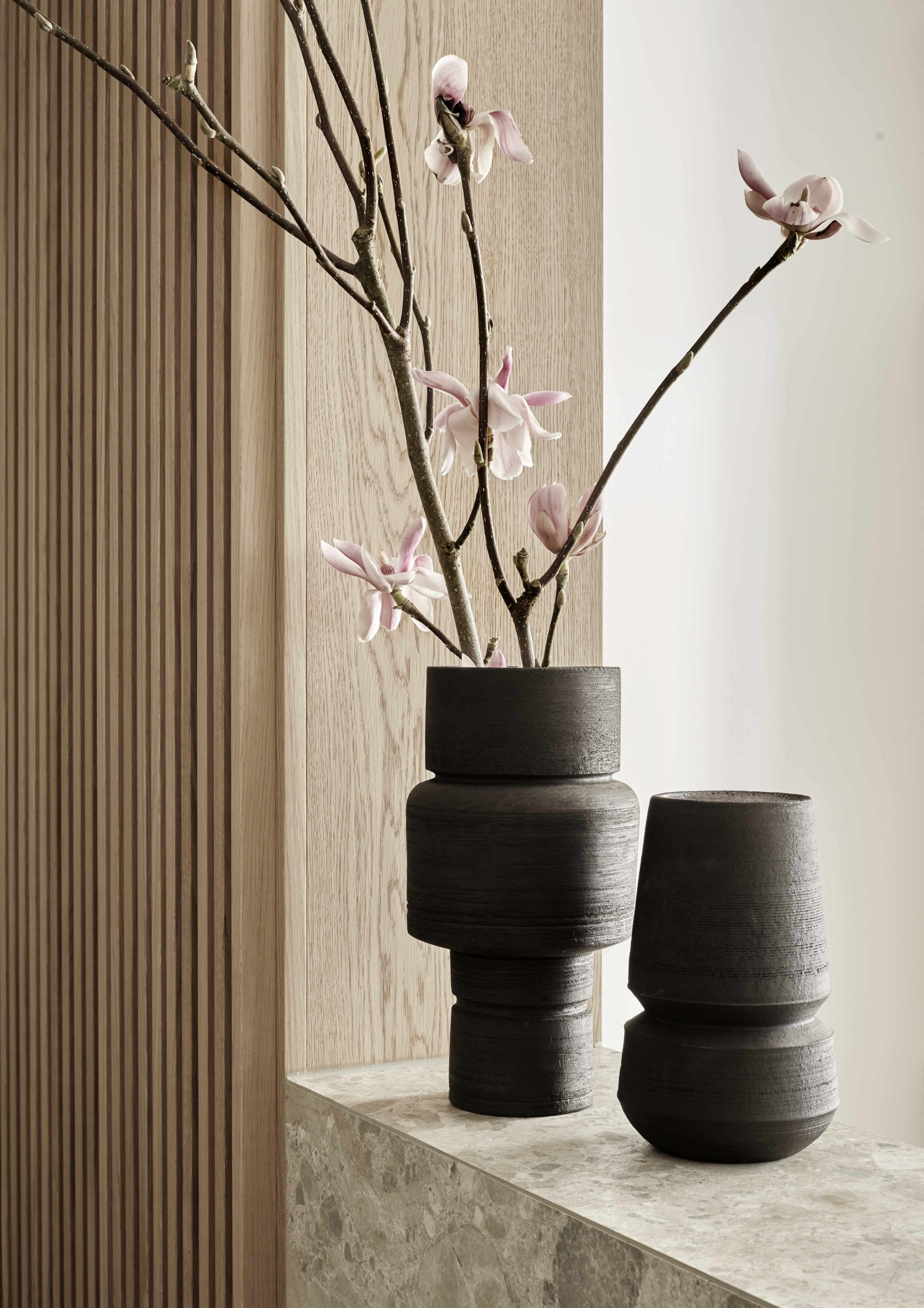 PEAM Praxis Detailfoto Magnolienzweig in schwarzen Keramik Vasen von Markus J. Freidl auf mit Terrazzo Fliesen belegtem Anmeldungstresen Holzlamellen in Eiche
