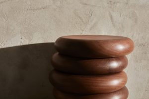 Skulpturaler Beistelltisch SLICE aus massivem Holz mit organisch gerundeter Form, arrangiert vor gespachtelter Kalkputzwand und Sichtestrichboden – Designobjekt mit handwerklichem Charakter.